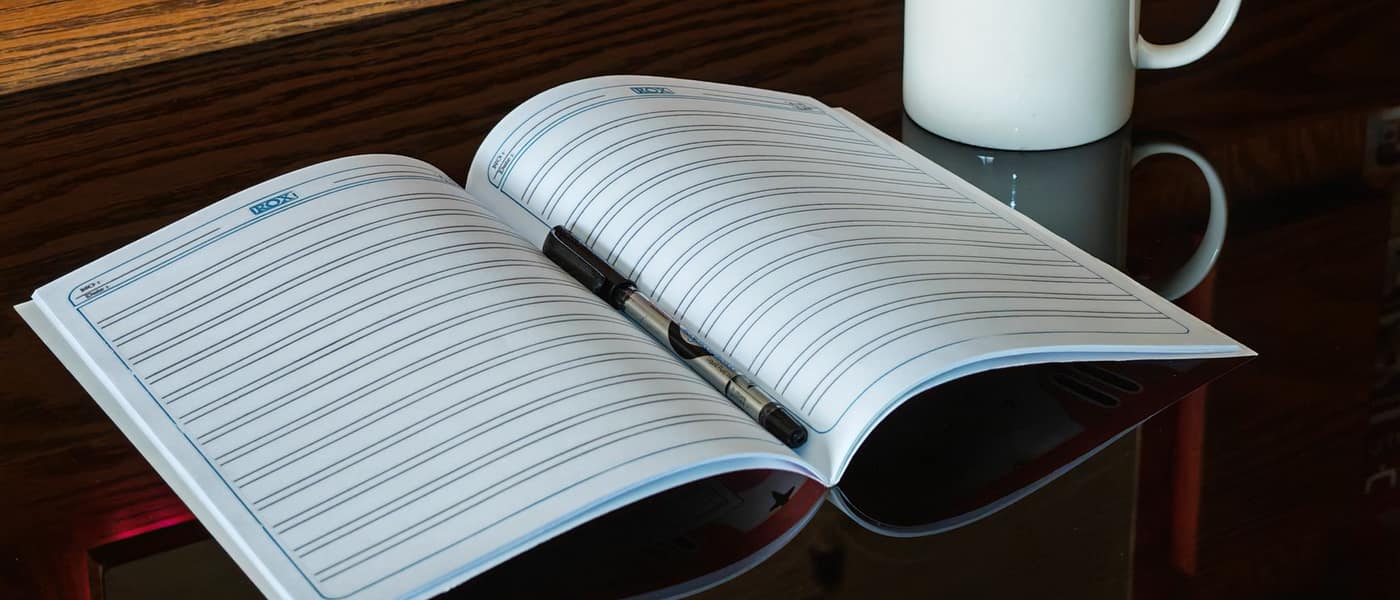 An open notebook with a pen beside a coffee mug on a table