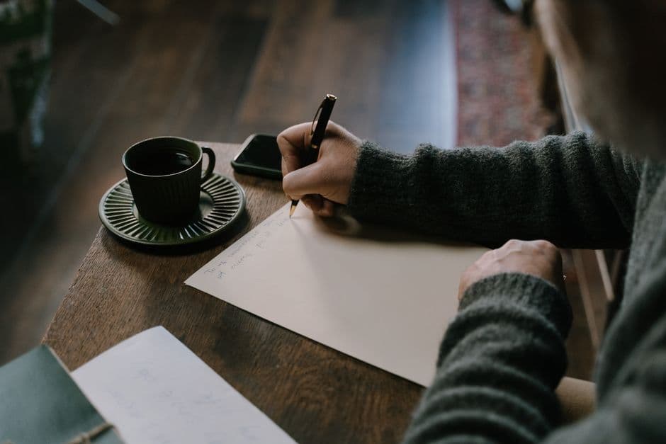 A person writing on paper at a table beside a cup of coffee