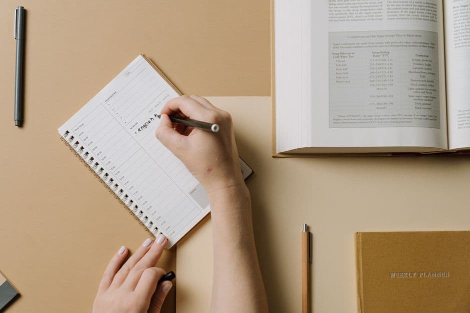 A person writing in a planner at a desk beside an open book