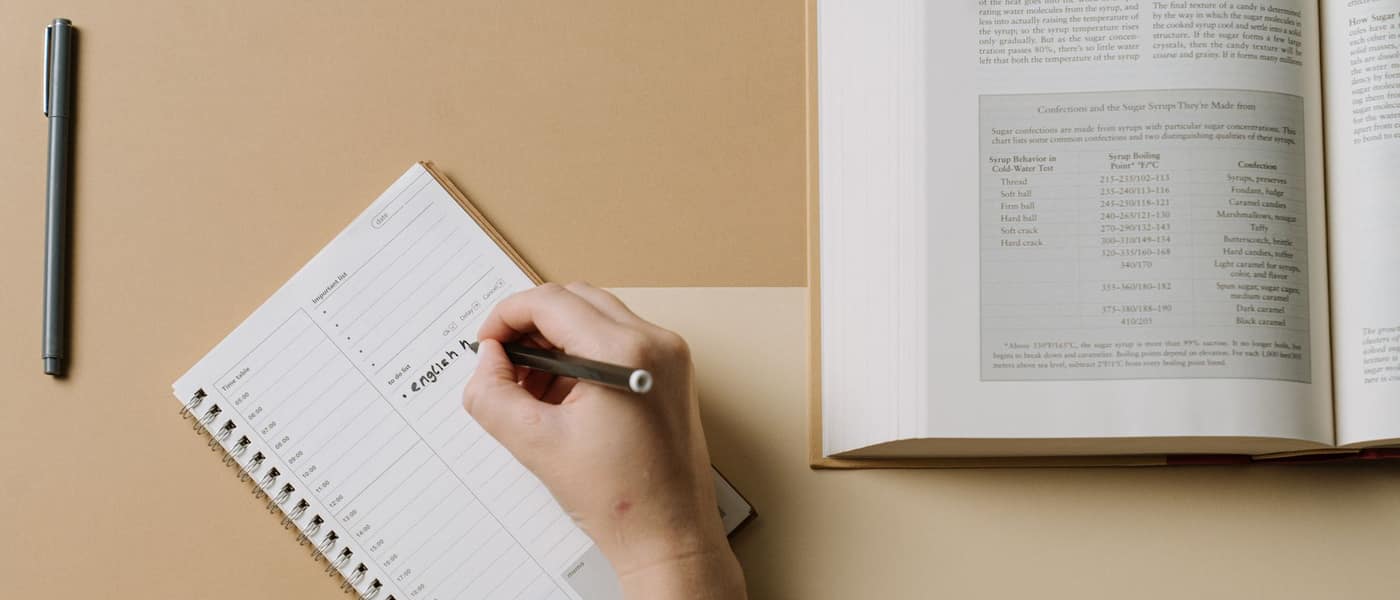 Hands writing in a planner at a desk beside an open book